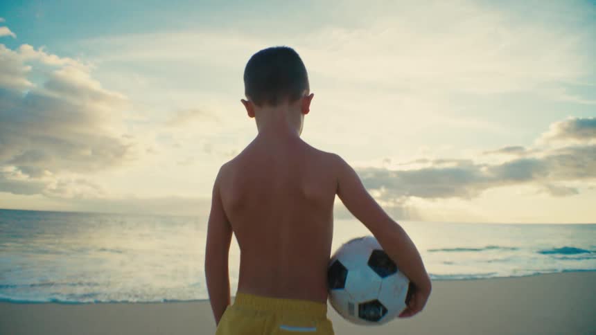 Boy without a shirt and wearing yellow shorts holding a soccer ball while standing on the sand at the beach