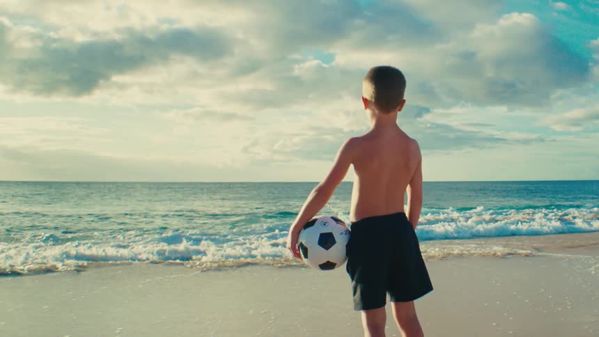 Back of young boy holding soccer ball standing at shoreline watching tide roll in