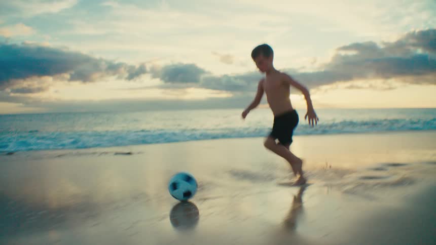 A young boy runs alone playing soccer on the beach