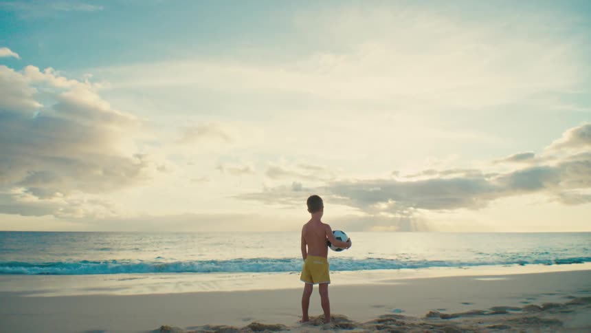 A young boy stands facing the beach holding a football in his hand