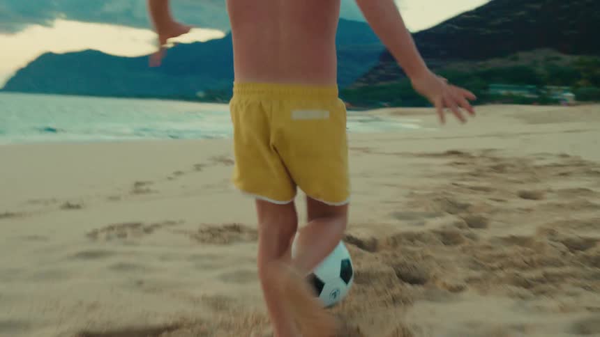 A kid wearing yellow trunks kicks a soccer ball on the beach