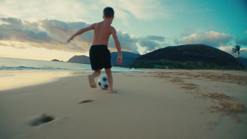 A young boy plays soccer barefoot on a sandy beach during sunset