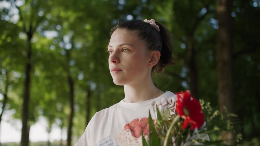 A young lady stands alone in the park looking bored while holding flowers but suddenly starts smiling as she stands under the trees