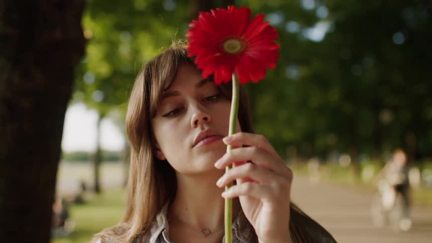 A woman with long brown hair holds a large red flower in front of her face standing outdoors in a park
