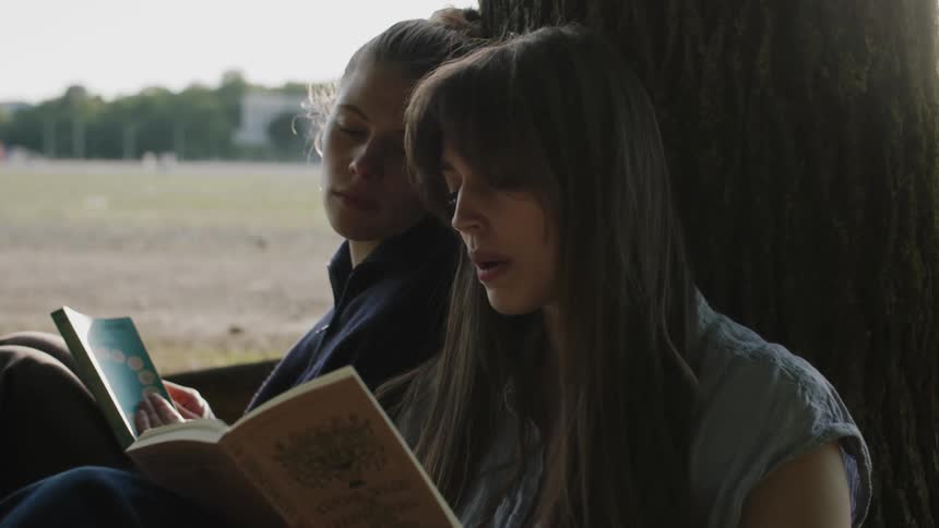 Two young women read books while sitting under a tree outdoors during daytime