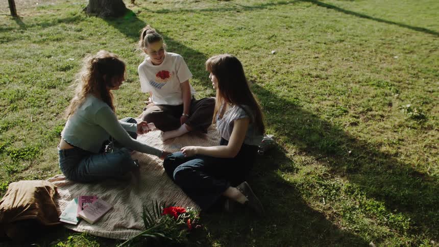 Three young women sit on a picnic blanket playing cards together in a sunny grassy park