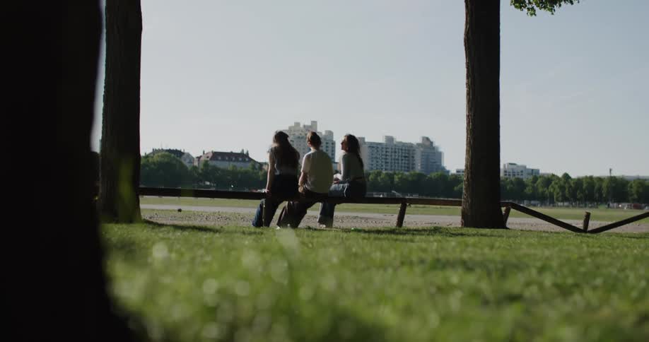 Three people sit on a bench in a park surrounded by trees with buildings visible in the background