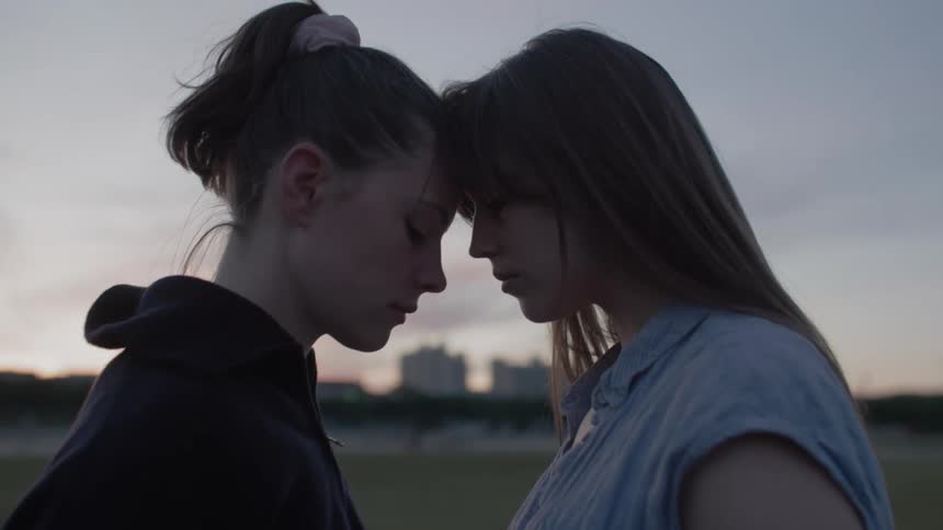 Two teenage women stand with their foreheads touching against a muted background with a faint sunset sky