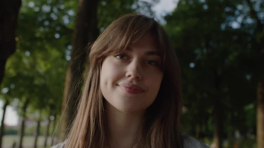 A woman with brown hair looks directly at the camera with a neutral expression standing outdoors among trees