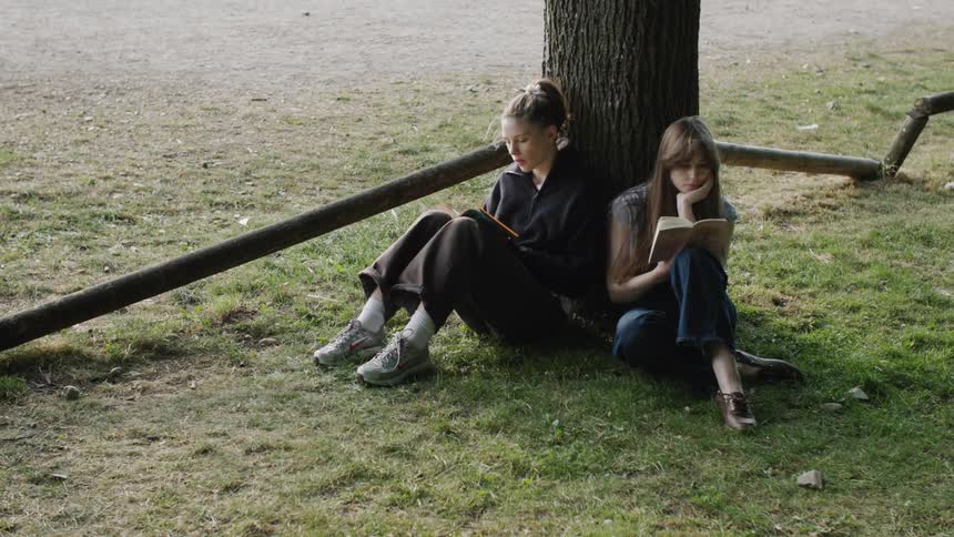 Two young women sit under a tree reading books enjoying a quiet moment in a park