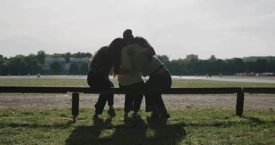 Three friends sit on a bench in the park hugging each other at midday