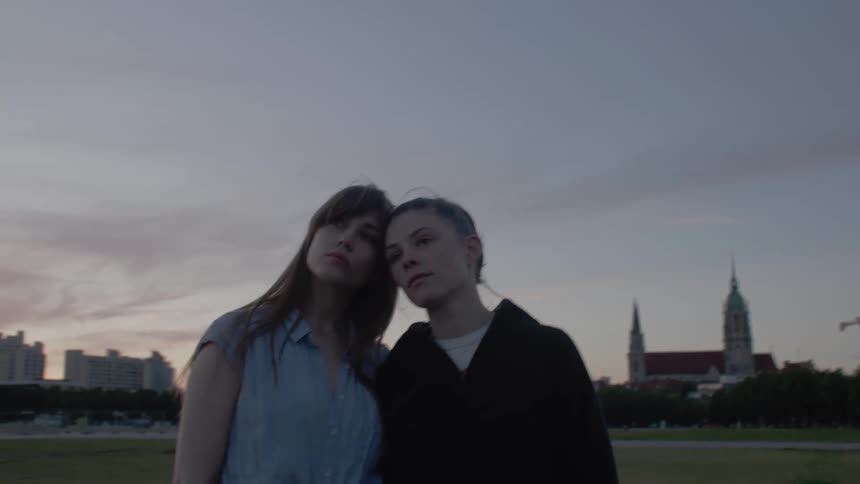 Two women stand close together at dusk one resting her head on the others shoulder with a city skyline silhouetted against a colorful sunset sky