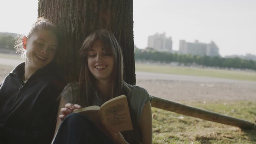 Two smiling women sitting under a tree reading a book