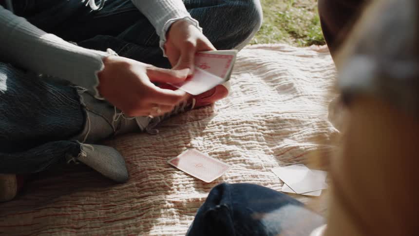 Three friends sit on the ground in a park playing a card game at midday
