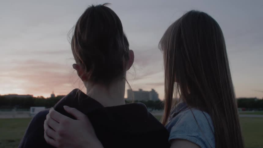 Two teenage girls with their backs to the camera stand close together one with an arm around the other watching the sunset over a grassy field with trees and buildings in the distance