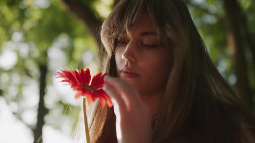A woman with long brown hair gently smells a red gerbera daisy in a sunlit outdoor setting