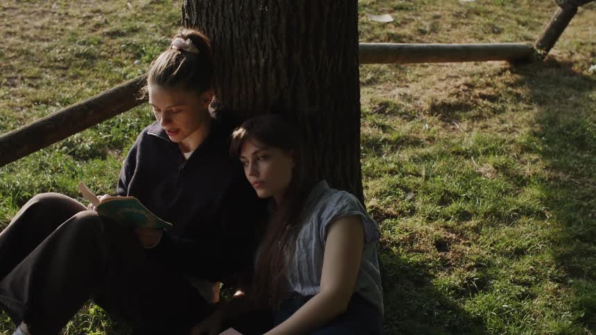 Two young women sit on grass leaning against a tree in a sunny outdoor setting with one reading a book
