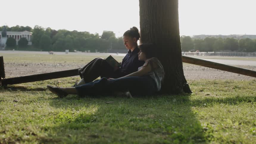 Two people relax under a tree in a sunlit park sharing a quiet peaceful moment