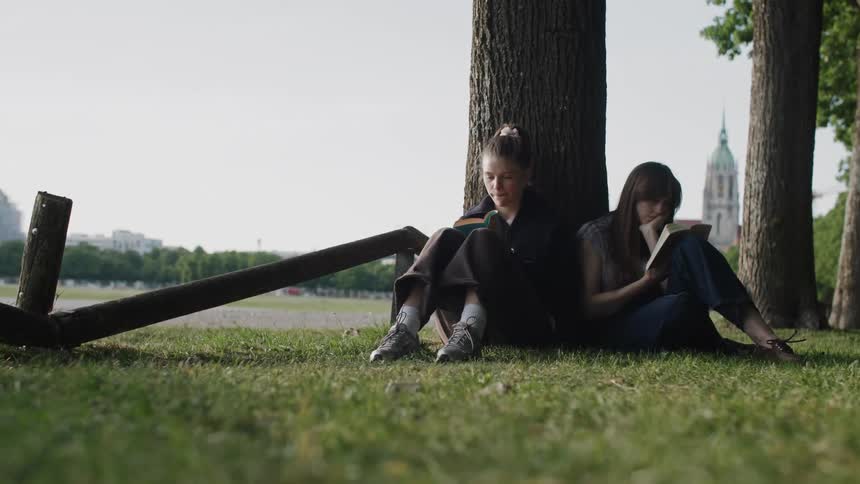 Two young women are sitting under a tree in a park peacefully reading books