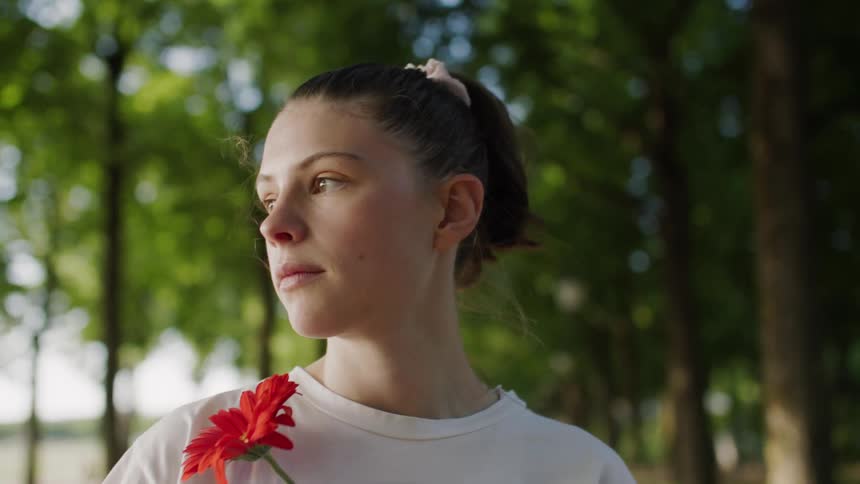 Woman holds a red flower while standing in the woods near a lake on a summer day