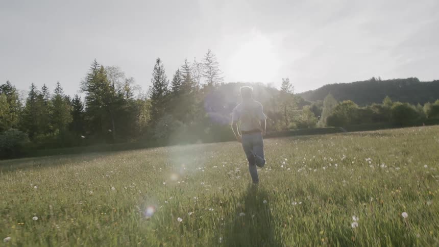 A man runs joyfully through a sunlit field surrounded by trees and hills