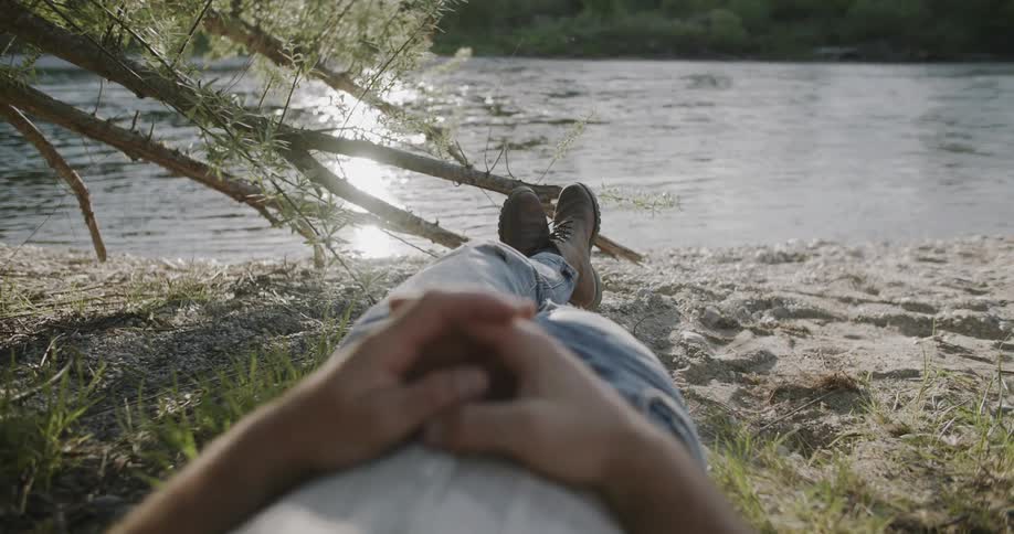 Person relaxing by a riverbank lying on the sand with folded hands
