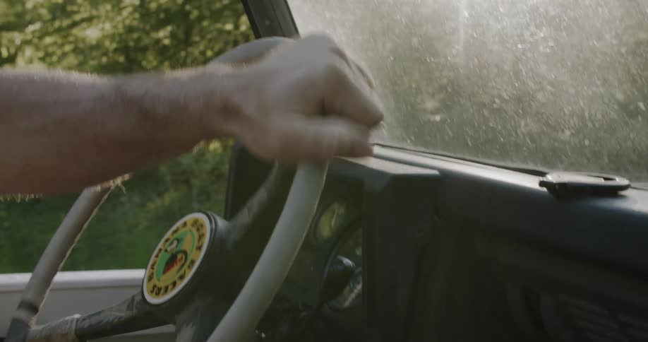 A mans arm and hand are visible on a white steering wheel inside a vehicle with foliage outside the windshield