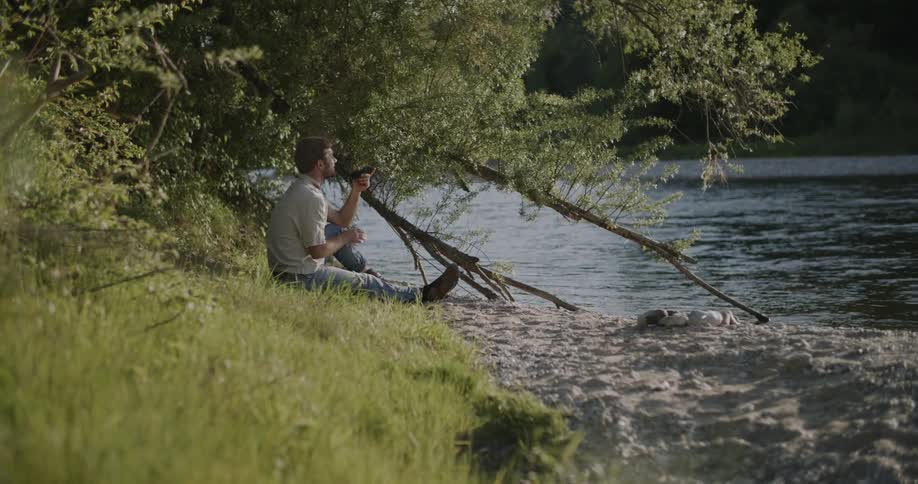 The man is sitting by the riverbed and talking on his phone
