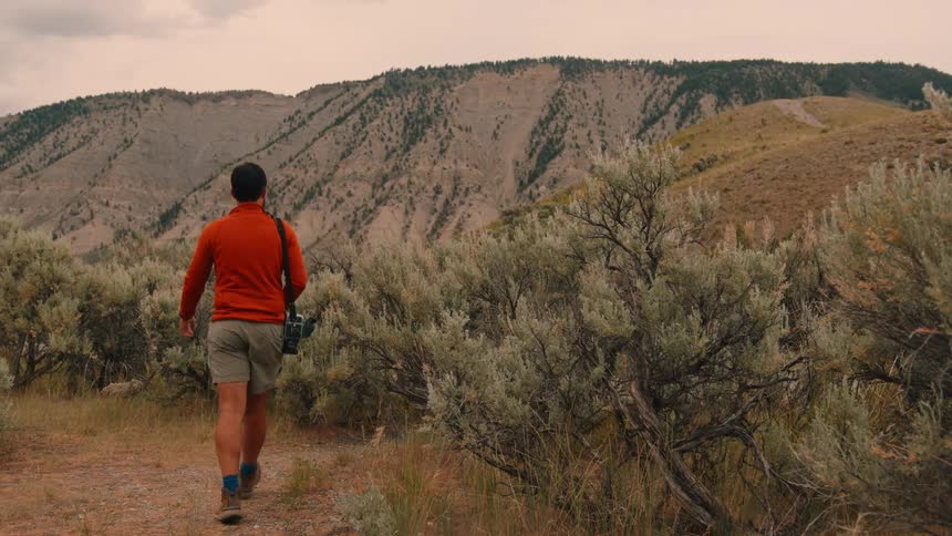 A man walks through a dry mountain landscape carrying a camera