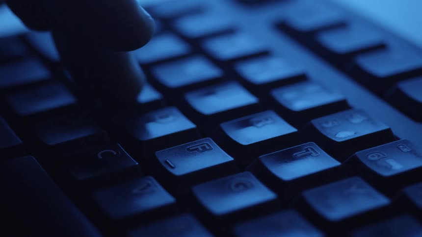 Close up of a computer keyboard as a person types in a blue lit room
