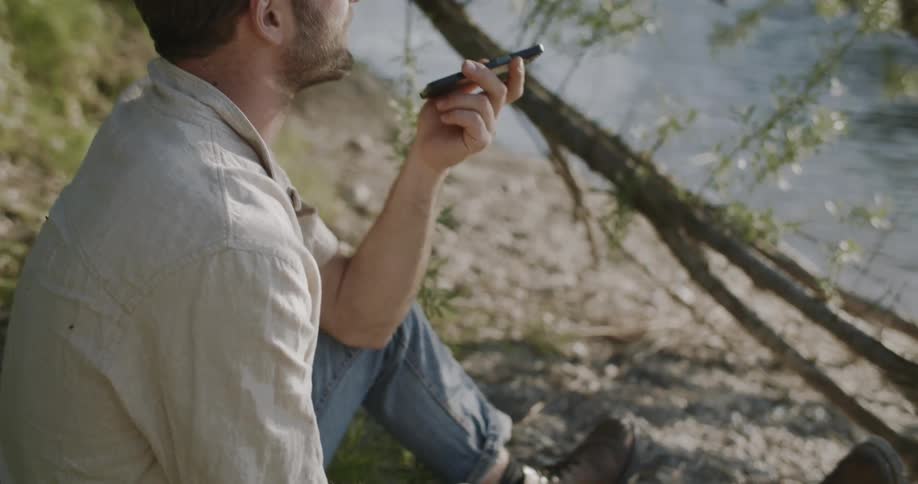 A man sits and talks on his phone by a tranquil river ending on the sun sparkling across the waters surface