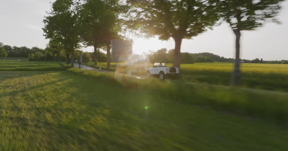 A white convertible drives along a narrow country road flanked by tall trees and green fields during a golden sunset