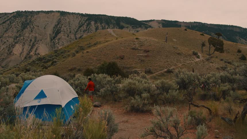 A person walks on top of a mountain away from a tent surrounded by grass all around at midday