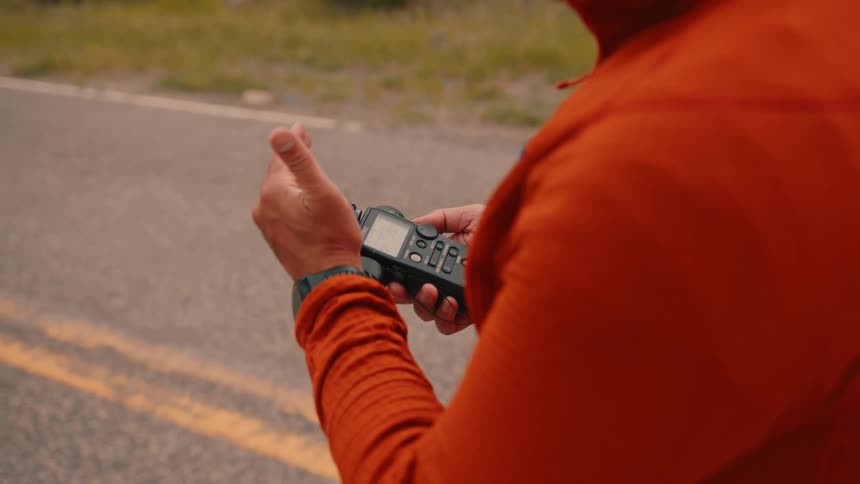 A man wearing an orange shirt stands in the middle of the road at midday holding a professionalgrade light meter
