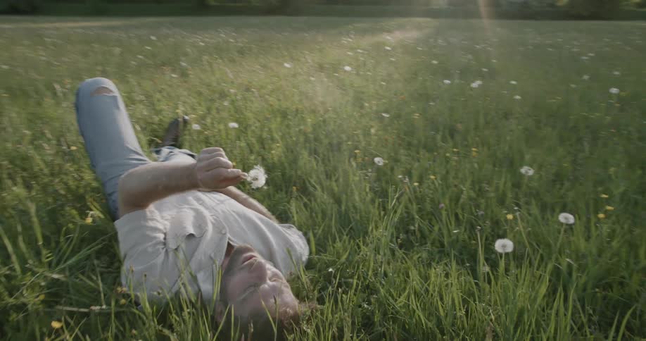 A man lies in a grassy field holding a dandelion in one hand and blowing its seeds into the air