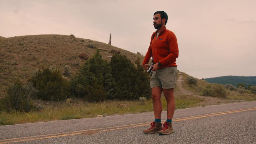 A man stands on a rural road photographing the ground in a mountainous landscape