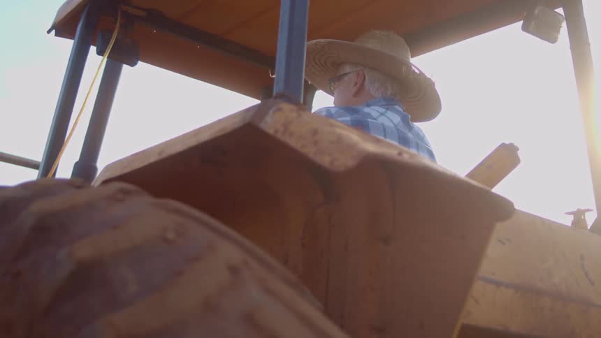 An elderly man in a straw hat and plaid shirt drives a yellow tractor looking away from the camera