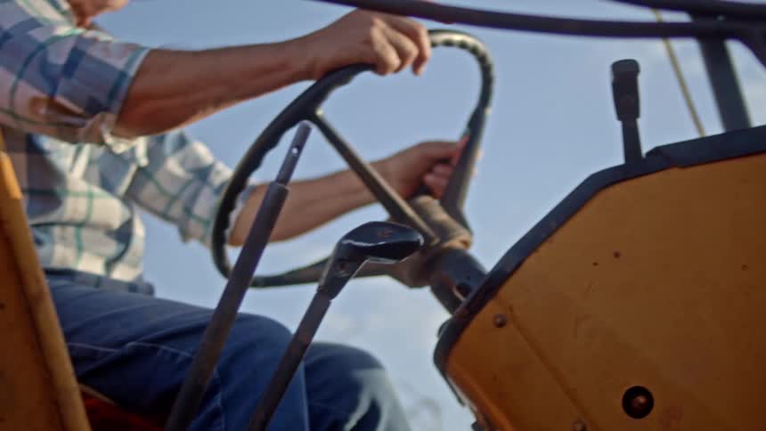 A persons hands grip the steering wheel of a yellow tractor with a bright sky in the background