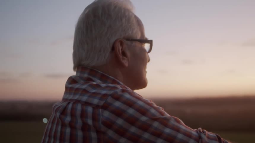 An elderly person wearing a plaid shirt stands outdoors at sunset gazing toward a sunlit field bordered by trees