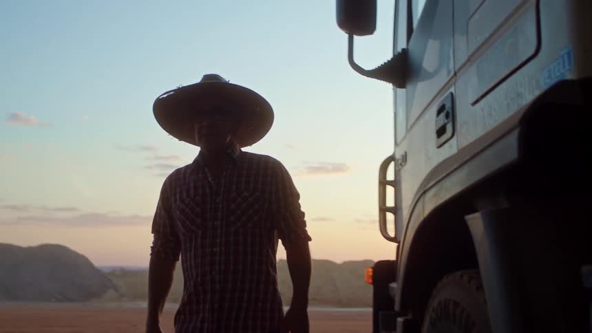 A farmer wearing a sun hat walks past a truck into a warehouse at midday