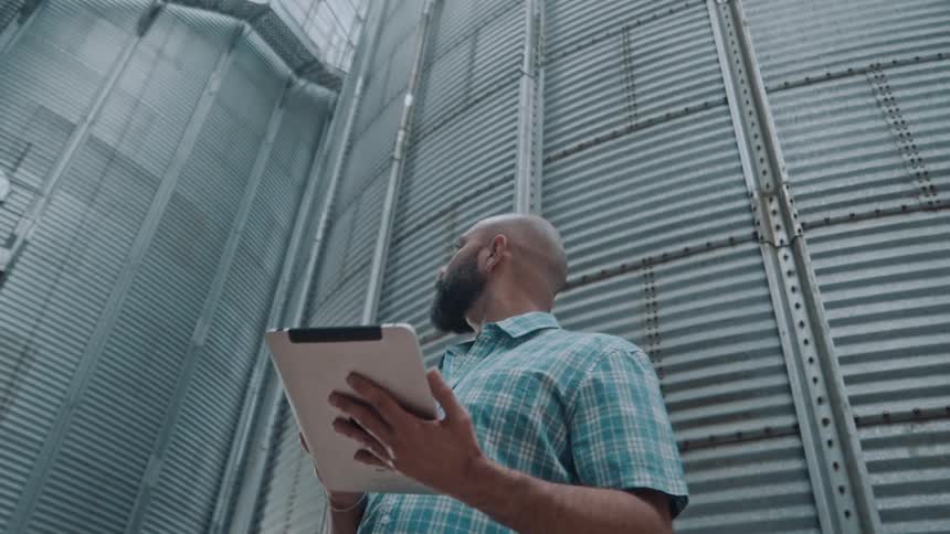 A man with a beard wearing a plaid shirt holds a white tablet and looks towards a large corrugated metal silo