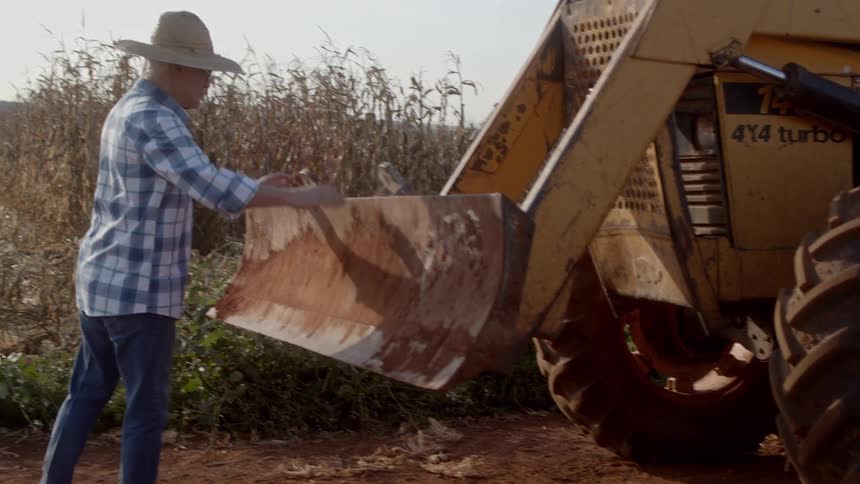 A farmer in a plaid shirt and jeans inspects the bucket of a yellow backhoe loader on a dirt road near a field