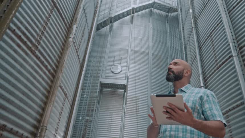 A man with a beard and a plaid shirt holds a tablet and looks upward while standing between two tall corrugated metal structures in a grain silo or storage area
