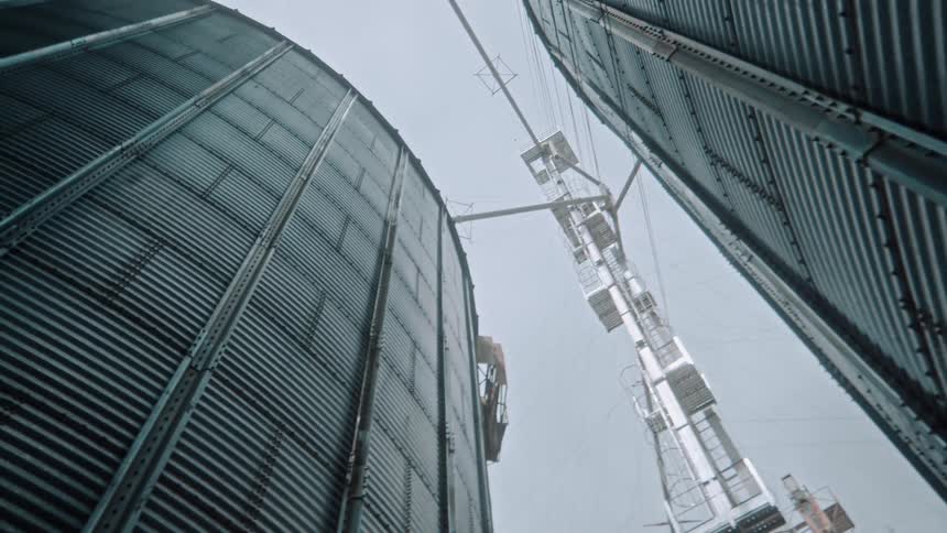 A lowangle tracking shot moves between towering corrugated metal grain silos looking up at the complex machinery and overcast sky above