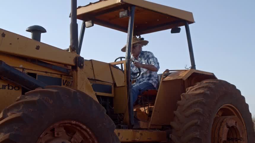 An elderly farmer wearing a straw hat is starting his tractor while at the corn field