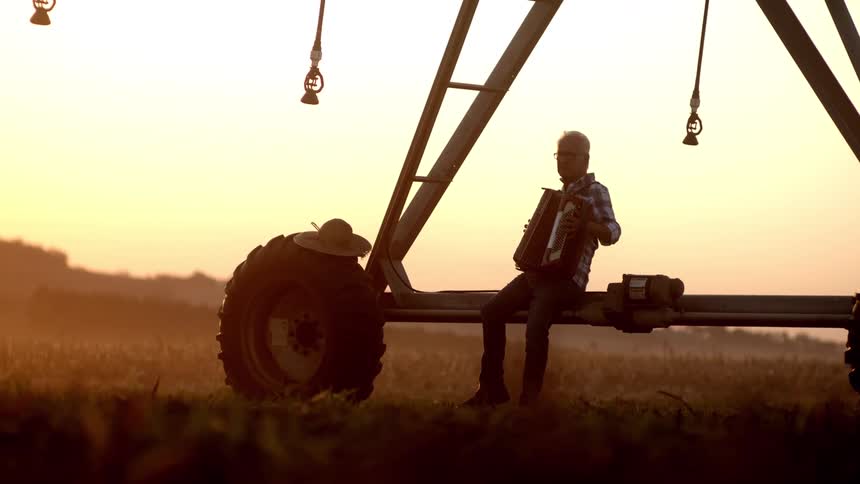 Elderly man sitting on farm machinery outdoors and playing harmonica