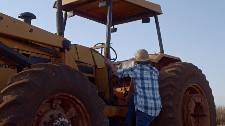 A farmer in a plaid shirt and straw hat climbs onto a large dusty tractor under a clear blue sky