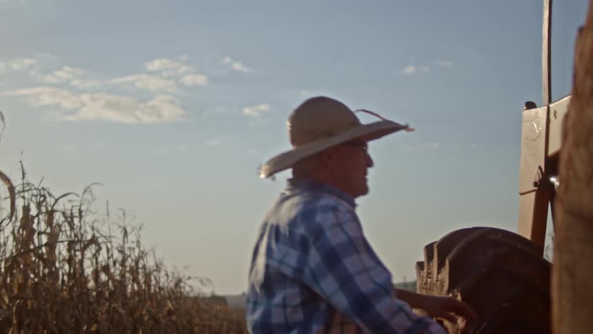 A man in a straw hat and plaid shirt steps out of a tractor to inspect the front with dry corn stalks in the foreground under a bright sky