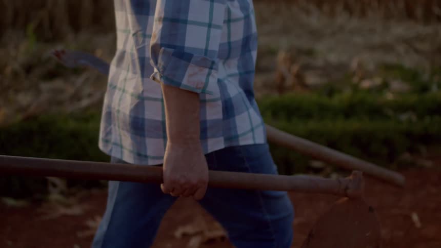 Male farmer carries two farm tools while walking on a farmland during daytime