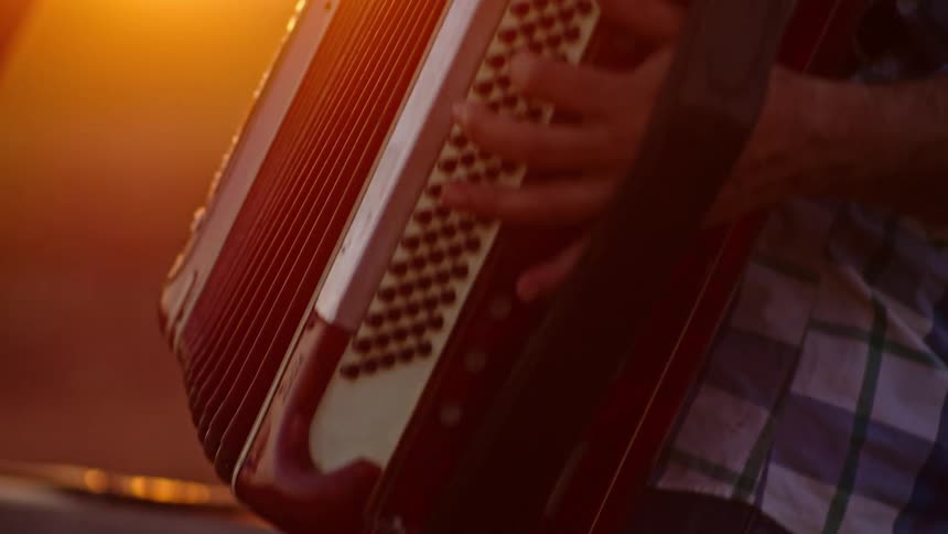 An older person wearing a plaid shirt plays an accordion outdoors at sunset